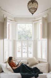 Junge Frau entspannt sich auf dem Wohnzimmersofa und liest ein Buch im Erkerfenster - FSIF05253