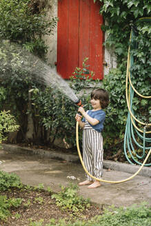 Smiling boy watering plant with garden hose while standing on footpath at back yard - ALBF01571