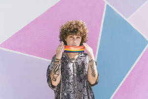 Young woman holding multi colored face mask while standing against wall - DAMF00488