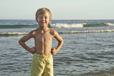 Lächelnder Junge mit Hand auf der Hüfte, der am Strand am Meer steht - VEGF02940