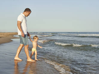 Junge hält die Hand seines Vaters beim Spaziergang am Strand - VEGF02941