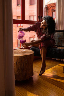 Ethnische lateinamerikanische Frau, die zu Hause am Fenster sitzend einen gesunden Kefir- und Pitaya-Smoothie genießt - ADSF15842