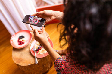 Von oben abgeschnittene unerkennbare Frau, die mit dem Handy beim Essen ein gesundes Frühstück mit Mandelmehlpfannkuchen mit Beeren und Honig fotografiert - ADSF15851