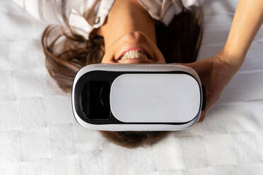 Von oben glückliche junge Frau mit VR-Headset, die lächelnd auf dem Bett liegt und den Cyberspace zu Hause erkundet - ADSF15854