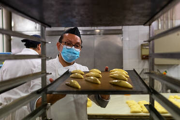 Männlicher Koch in Uniform und mit medizinischer Maske bei der Zubereitung von leckerem Gebäck in der Küche einer Bäckerei während einer Coronavirus-Epidemie - ADSF15855