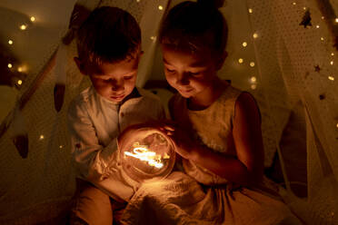 Geschwister, die ein Licht halten, während sie zu Weihnachten im Zimmer sitzen - GMLF00649