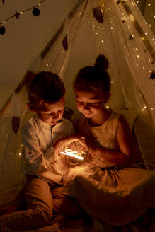 Bruder und Schwester sitzen im Zimmer und halten ein Weihnachtslicht - GMLF00650