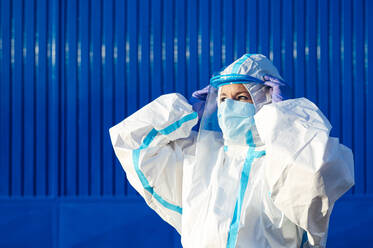 Nurse wearing face shield while standing against blue wall on sunny day - PGF00119