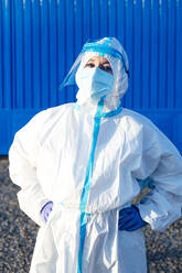 Female nurse with hand on hip standing against blue wall on sunny day - PGF00120