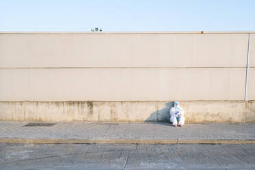 Nurse wearing protective suit sitting on footpath against wall - PGF00125