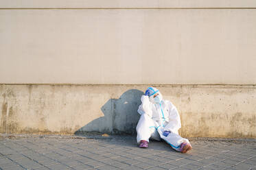 Female nurse sitting with head in hands on footpath against wall - PGF00126