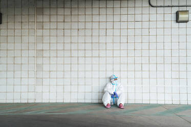 Female nurse wearing protective suit with sitting against tiled wall - PGF00130