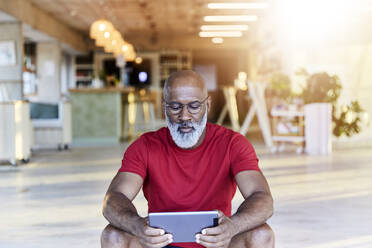 Älterer Mann in rotem T-Shirt, der auf einem Dach sitzend ein digitales Tablet benutzt - FMKF06412