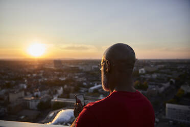 Kahlköpfiger Mann fotografiert mit seinem Smartphone von der Terrasse aus den Sonnenuntergang über der Stadt - FMKF06421