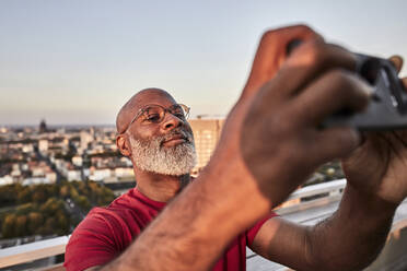 Bärtiger reifer Mann, der sein Smartphone benutzt, um den Sonnenuntergang von einem Hausdach in der Stadt zu fotografieren - FMKF06423