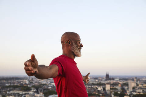 Happy completely bald man standing with arms outstretched on building terrace in city during sunset stock photo