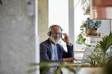 Lächelnder Geschäftsmann hört über Kopfhörer zu, während er mit seinem Laptop am Tisch sitzt und von zu Hause aus arbeitet - FMKF06433