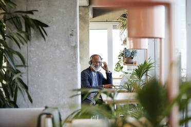 Smiling businessman listening through headphones while sitting with laptop at table and working from home - FMKF06434