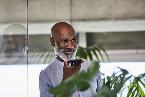 Lächelnder reifer Geschäftsmann, der einen Anruf über sein Smartphone entgegennimmt, während er sich an eine Glaswand lehnt - FMKF06437