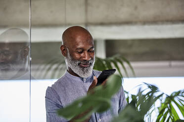 Smiling mature freelancer listening to call through smart phone while leaning on glass wall - FMKF06438