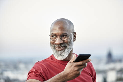 Smiling bald man talking through smart phone while looking away at building terrace during sunset - FMKF06448