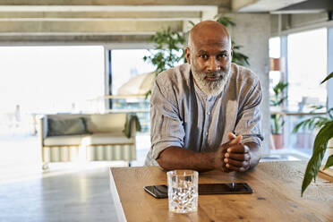 Confident bald man with hands clasped leaning on table at rooftop - FMKF06453