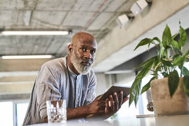 Reifer Mann mit Glatze, der ein digitales Tablet hält, während er sich auf einen Tisch stützt und auf eine Gebäudeterrasse blickt - FMKF06456