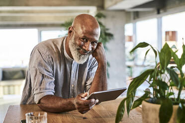 Lächelnder reifer Mann, der ein digitales Tablet hält, während er sich auf einen Tisch auf einer Gebäudeterrasse stützt - FMKF06457