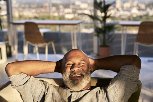 Lächelnder reifer Mann entspannt sich auf dem Sofa auf der Terrasse eines Gebäudes bei Sonnenuntergang - FMKF06461
