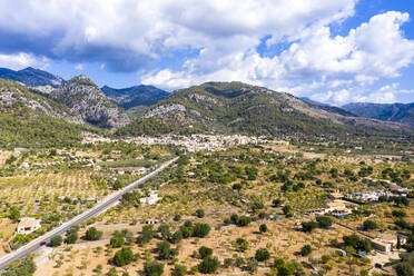 Luftaufnahme einer Straße in Richtung eines Dorfes in der Nähe einer Gebirgskette bei bewölktem Himmel, Caimari, Mallorca, Spanien - AMF08518