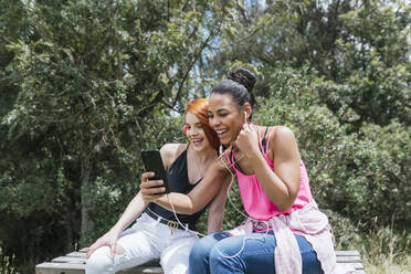 Cheerful young woman taking selfie with redhead female friend sitting at park on sunny day - DSIF00143