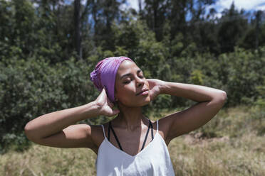 Junge Frau trägt lila Kopftuch, während sie mit geschlossenen Augen im Park an einem sonnigen Tag steht - DSIF00144
