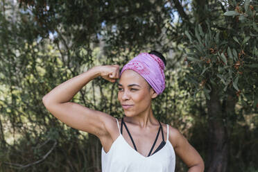Young woman with purple bandana flexing bicep muscles while standing against plants at park - DSIF00146