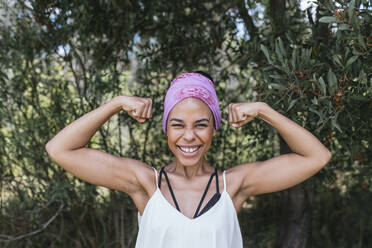 Happy woman with purple bandana flexing muscles while standing against plants at park - DSIF00147