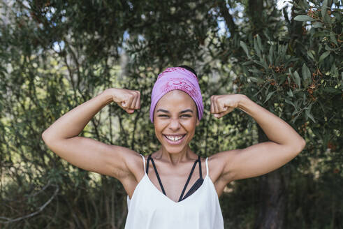 Glückliche Frau mit lila Kopftuch, die ihre Muskeln anspannt, während sie im Park vor Pflanzen steht - DSIF00147