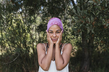 Young woman with hands on cheeks wearing purple bandana while standing against plants at park - DSIF00148