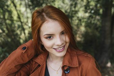 Lächelnde schöne junge rothaarige Frau mit brauner Jacke im Park - DSIF00151