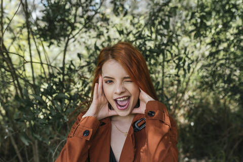 Playful young redhead woman winking while standing at park stock photo