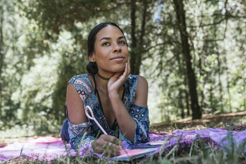 Thoughtful young woman looking away while lying on picnic blanket with diary at park stock photo