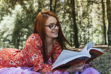 Smiling young beautiful redhead woman reading book while lying on picnic blanket with diary at park - DSIF00156