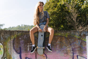 Nachdenkliche blonde Frau mit Skateboard, die auf einer Mauer in einem Park sitzt - FMOF01151