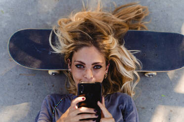 Blonde Frau benutzt ihr Smartphone, während sie auf einem Skateboard über der Straße liegt - FMOF01153