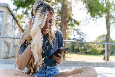 Young blond woman using smart phone while enjoying listening music through headphones at skateboard park - FMOF01156