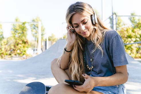 Junge blonde Frau hört Musik über Kopfhörer, während sie ihr Smartphone im Skateboard-Park benutzt - FMOF01157