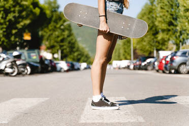 Frau, die ein Skateboard hält, während sie auf einem Zebrastreifen in der Stadt steht - FMOF01159