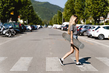 Junge blonde Frau hält ein Skateboard, während sie über einen Zebrastreifen in der Stadt läuft - FMOF01160