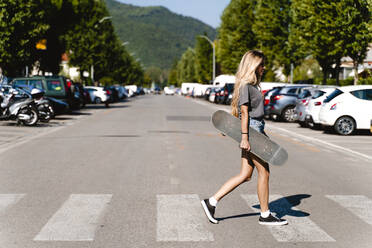 Young blond woman holding skateboard while walking on zebra crossing in city - FMOF01160