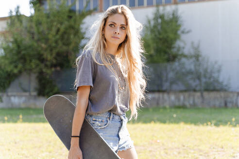 Schöne junge blonde Frau hält Skateboard, während sie in der Stadt an einem sonnigen Tag wegschaut - FMOF01163