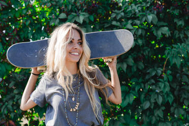 Lächelnde blonde Frau, die ein Skateboard hält und vor einer grünen Pflanze wegschaut - FMOF01166