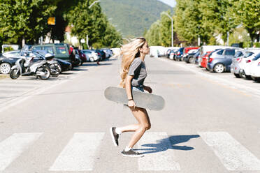 Junge blonde Frau hält Skateboard, während sie über einen Zebrastreifen in der Stadt läuft - FMOF01168